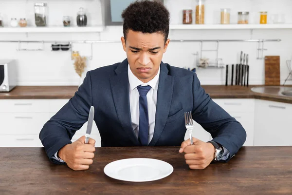 Frustrated african american man holding cutlery and looking at empty plate — Stock Photo