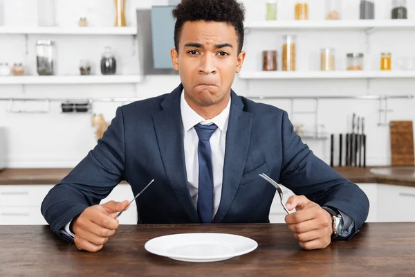 Hombre afroamericano trastornado sosteniendo cubiertos y mirando el plato vacío - foto de stock