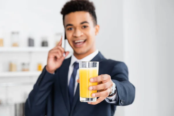 Стакан апельсинового сока в руке счастливого африканского бизнесмена, разговаривающего по смартфону на размытом фоне — стоковое фото