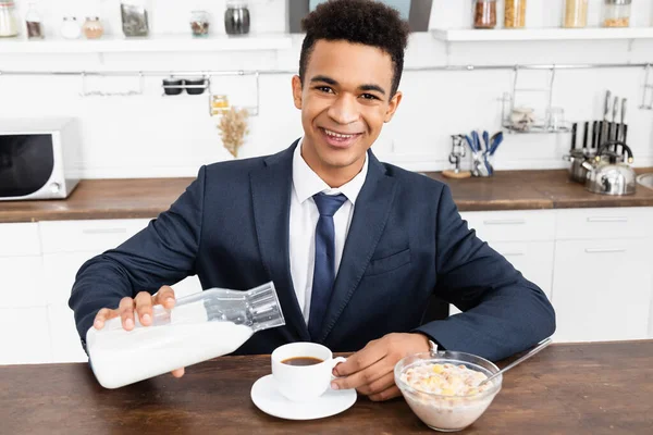 Happy african american businessman pouring milk in coffee near bowl with corn flakes — Stock Photo
