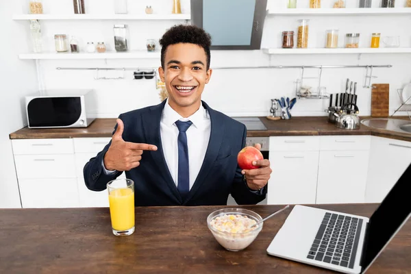 Feliz hombre de negocios afroamericano apuntando con el dedo a la manzana cerca del vaso de jugo de naranja, hojuelas de maíz y portátil - foto de stock