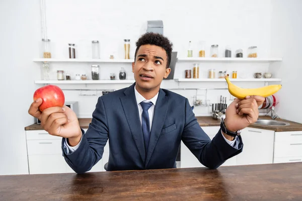 Puzzled african american businessman choosing between apple and banana — Stock Photo