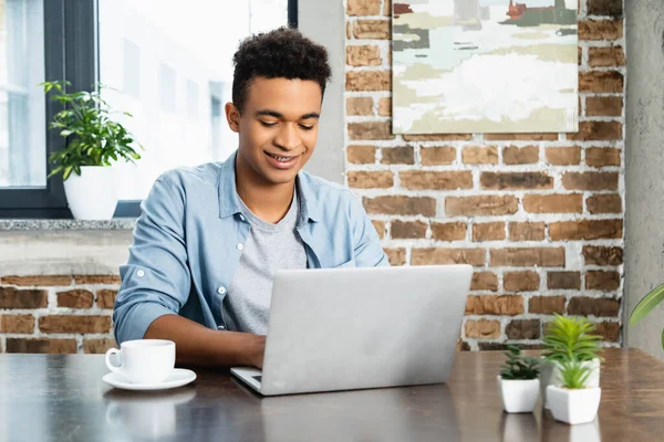 Pleased african american man using laptop near cup on desk — Stock Photo