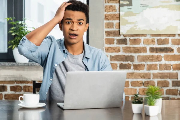 Sorprendido hombre afroamericano mirando la cámara cerca de la computadora portátil en el escritorio - foto de stock