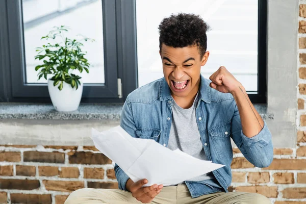 Excité homme afro-américain avec poing serré tenant des documents tout en célébrant le triomphe — Photo de stock