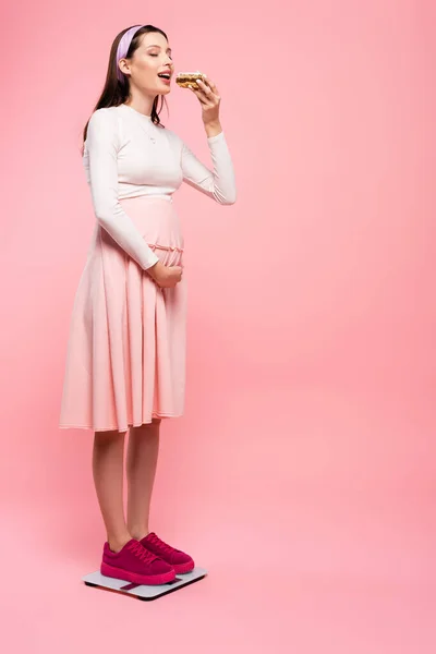 Young pretty pregnant woman eating cake on scales isolated on pink — Stock Photo