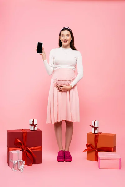 Улыбающаяся молодая красивая беременная женщина со смартфоном рядом с подарками, изолированными на розовый — стоковое фото