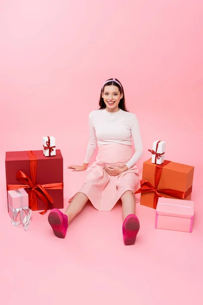 Усміхнена молода красива вагітна жінка сидить на підлозі з подарунками ізольована на рожевому — стокове фото