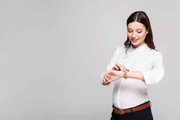 Усміхнена молода вагітна бізнес-леді дивиться на наручний годинник ізольовано на сірому — стокове фото