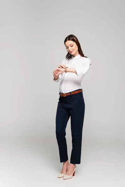 Smiling young pregnant businesswoman looking at wristwatch isolated on grey — Stock Photo