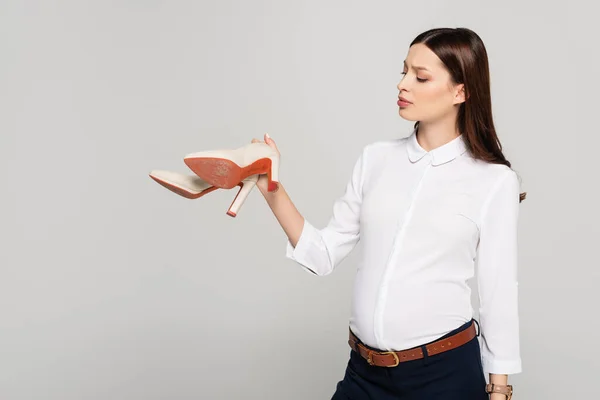 Joven embarazada mujer de negocios sosteniendo zapatos de tacón aislados en gris - foto de stock