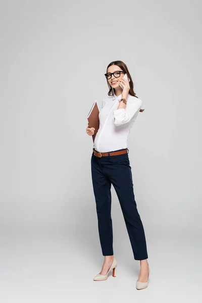 Smiling young pregnant businesswoman in glasses with folder talking on smartphone isolated on grey — Stock Photo