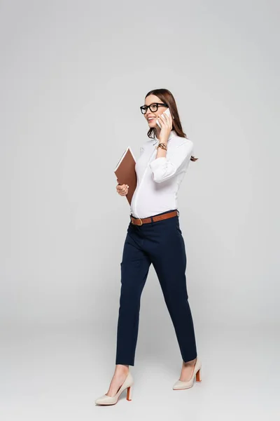 Smiling young pregnant businesswoman in glasses with folder talking on smartphone isolated on grey — Stock Photo