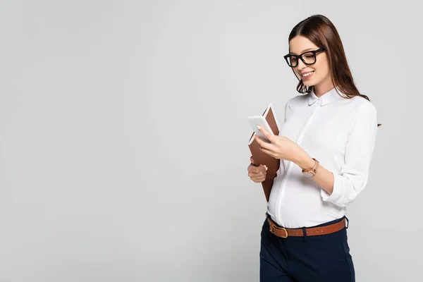 Улыбающаяся молодая беременная деловая женщина в очках с папкой и смартфоном, изолированными на сером — стоковое фото