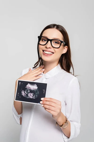 Sonriente joven embarazada mujer de negocios en gafas sosteniendo ultrasonido escaneado aislado en gris - foto de stock