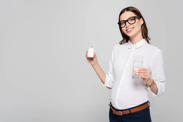 Sorridente jovem empresária grávida em copos com copo de água e vitaminas pré-natais isolado em cinza — Fotografia de Stock