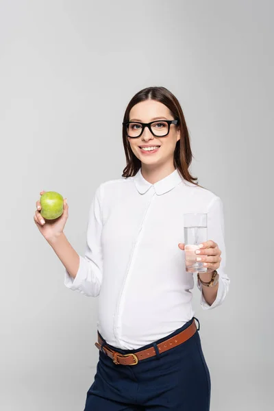 Sorridente giovane donna d'affari incinta in bicchieri con bicchiere d'acqua e mela isolata su grigio — Foto stock