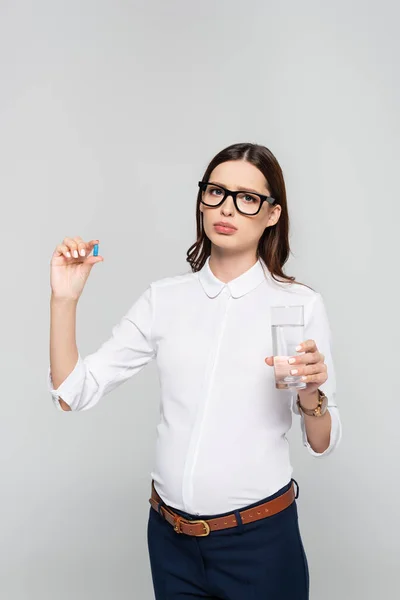 Triste joven empresaria embarazada en vasos con vaso de agua y píldora prenatal aislada en gris - foto de stock