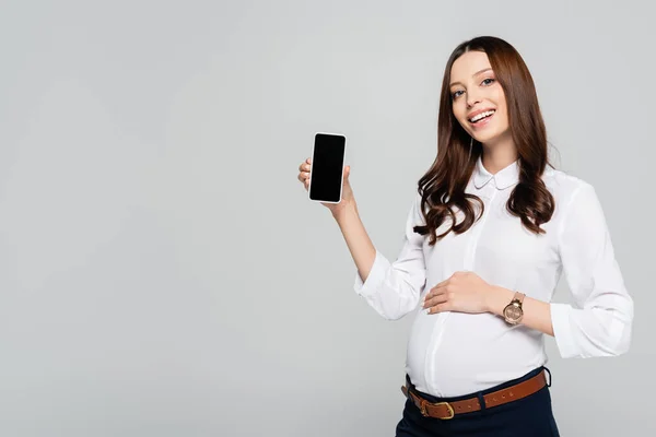 Улыбающаяся молодая беременная деловая женщина представляет смартфон, изолированный на сером — стоковое фото