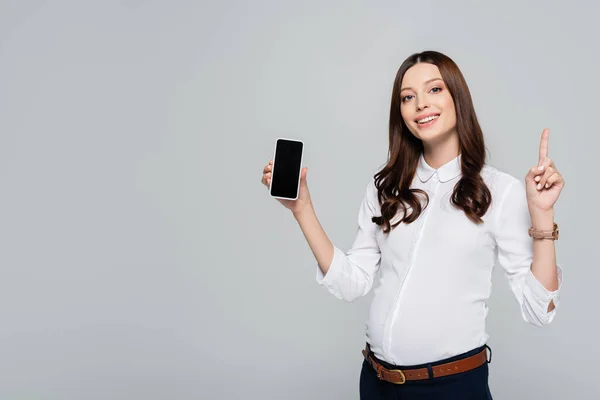 Sonriente joven mujer de negocios embarazada apuntando hacia arriba y presentando teléfono inteligente aislado en gris - foto de stock