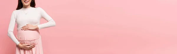 Vista ritagliata di felice giovane bella donna incinta toccando pancia isolata su rosa, banner — Foto stock