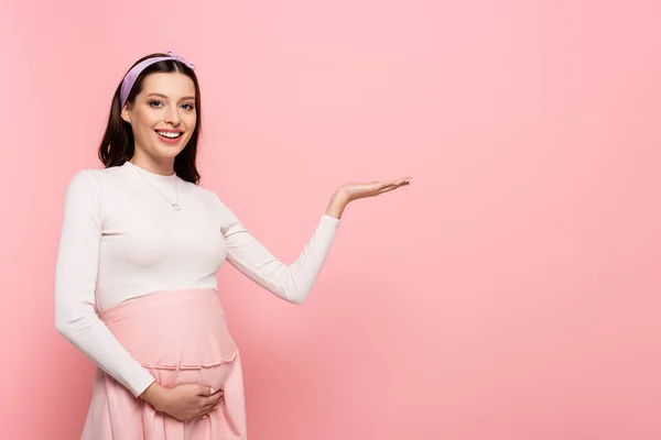 Heureux jeune jolie femme enceinte toucher le ventre et pointant de côté isolé sur rose — Photo de stock