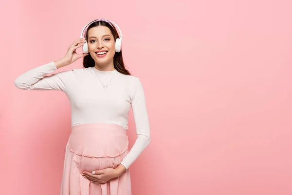 Heureux jeune jolie femme enceinte dans écouteurs toucher le ventre isolé sur rose — Photo de stock