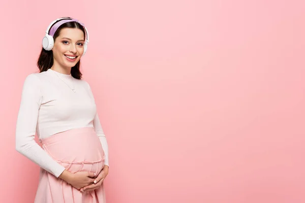 Щаслива молода красива вагітна жінка в навушниках, торкаючись живота, ізольована на рожевому — Stock Photo