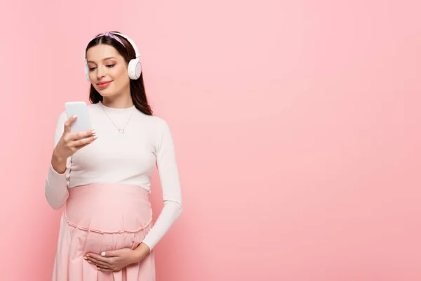 Happy young pretty pregnant woman in headphones using smartphone isolated on pink — Stock Photo