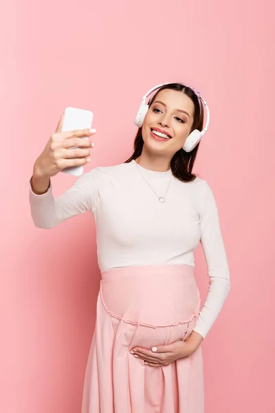 Счастливая молодая красивая беременная женщина в наушниках, используя смартфон изолирован на розовый — стоковое фото