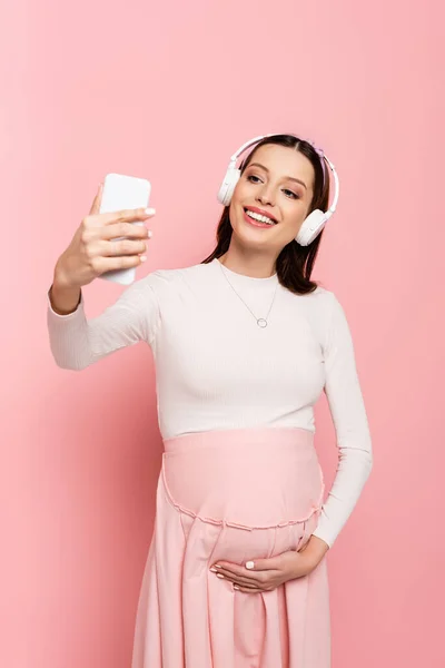 Heureux jeune jolie femme enceinte dans les écouteurs en utilisant smartphone isolé sur rose — Photo de stock