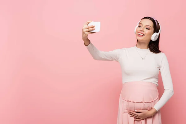 Heureux jeune jolie femme enceinte dans les écouteurs en utilisant smartphone isolé sur rose — Photo de stock