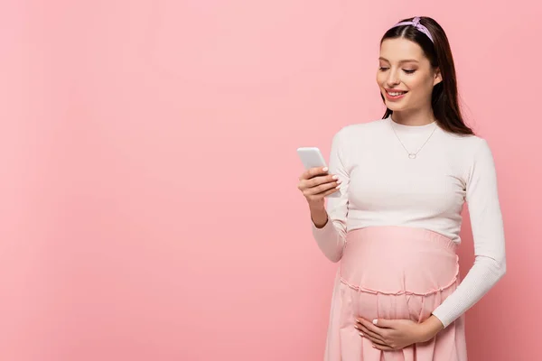 Счастливая молодая красивая беременная женщина с помощью смартфона изолированы на розовый — стоковое фото