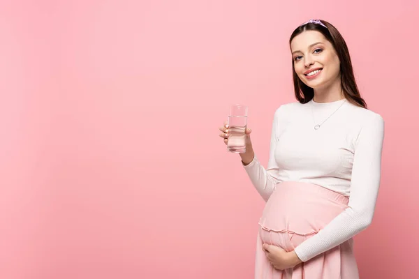Щаслива молода красива вагітна жінка зі склянкою води ізольована на рожевому — стокове фото
