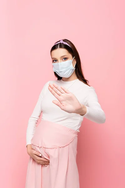 Молода вагітна жінка в медичній масці демонструє стоп- жест ізольовано на рожевому — стокове фото