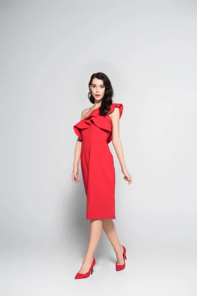 Brunette woman in red dress and heels looking at camera on grey background — Stock Photo