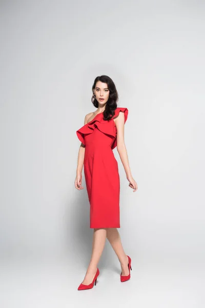 Brunette femme en robe rouge et talons regardant la caméra sur fond gris — Photo de stock