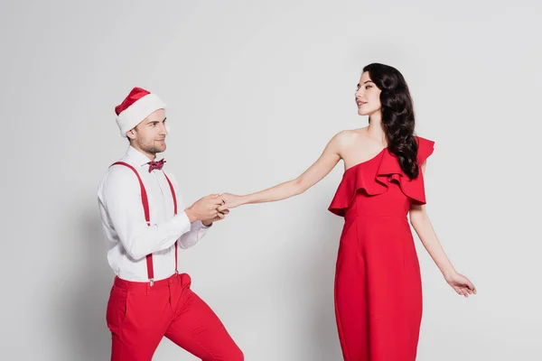 Young man in santa hat holding hand of woman in red dress on grey background — Stock Photo