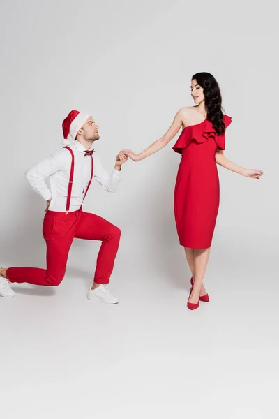 Man in santa hat standing on one knee and holding hand of smiling woman in red dress on grey background — Stock Photo