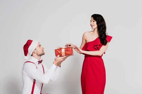 Man in santa hat holding present near smiling woman in red dress on grey background — Stock Photo
