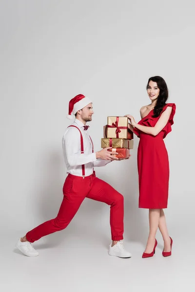 Frau in rotem Kleid und High Heels lächelt in die Kamera neben Mann mit Weihnachtsmütze mit Geschenken auf grauem Hintergrund — Stockfoto