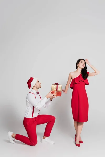 Homme en santa chapeau tenant des cadeaux près de femme souriante en robe rouge sur fond gris — Photo de stock