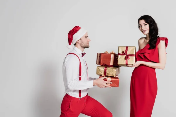Brunette femme souriant tout en tenant cadeau près de l'homme dans le chapeau de Père Noël avec des cadeaux sur fond gris — Photo de stock