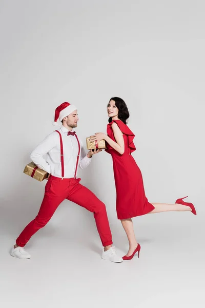 Lächelnde Frau in Kleid und High Heels hält Geschenk neben Mann mit Weihnachtsmütze versteckt Geschenk auf grauem Hintergrund — Stockfoto