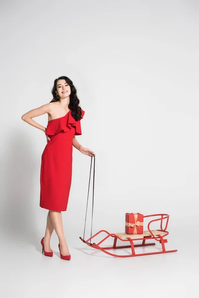 Улыбающаяся брюнетка в красном платье, стоящая возле саней с подарком на сером фоне — стоковое фото