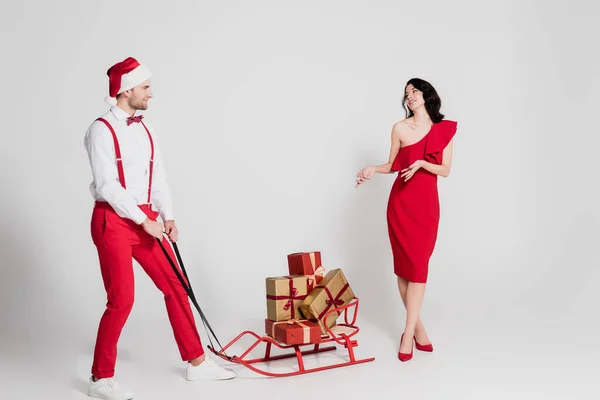 Homme au chapeau de Père Noël debout près du traîneau avec des cadeaux et femme souriante en robe sur fond gris — Photo de stock