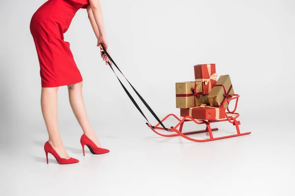 Vue recadrée de la femme en robe rouge et talons debout près des cadeaux sur traîneau sur fond gris — Photo de stock