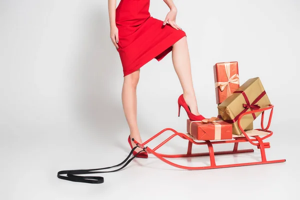 Vista recortada de la mujer en vestido rojo y tacones de pie cerca del trineo y regalos sobre fondo gris - foto de stock