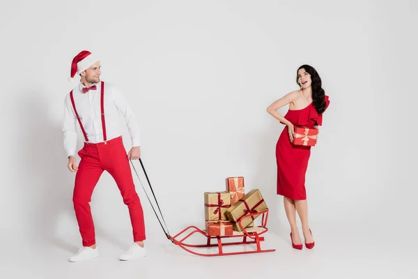 Homme souriant en santa chapeau tenant traîneau avec des cadeaux près de la femme en robe rouge sur fond gris — Photo de stock