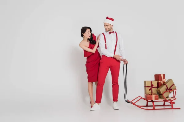 Aufgeregte Frau steht neben lächelndem Mann mit Weihnachtsmütze und deutet auf Schlitten mit Geschenken auf grauem Hintergrund — Stockfoto